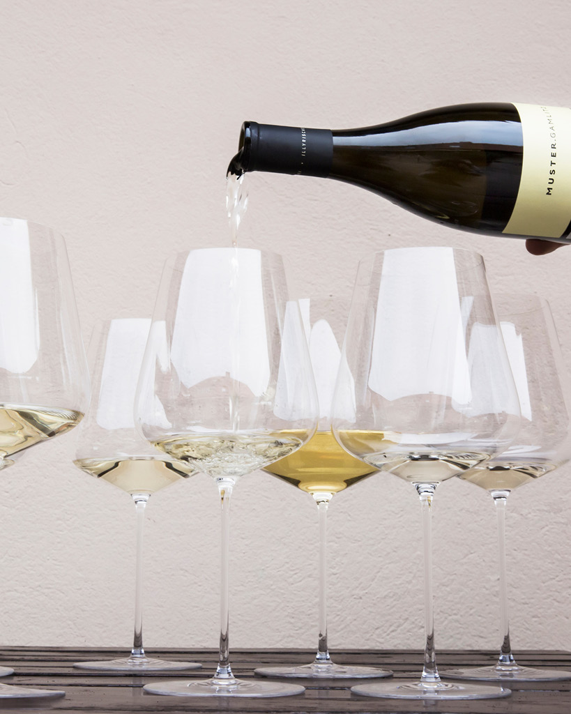 Weißwein wird aus einer Flasche in ein Weinglas gegossen, umgeben von weiteren gefüllten Weingläsern auf einem Holztisch vor einer hellen Wand