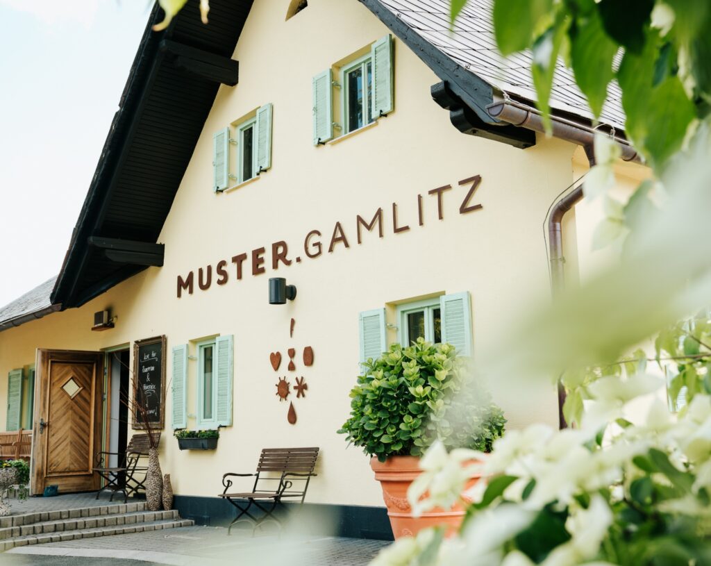 Hausfassade mit Schriftzug 'MUSTER.GAMLITZ', Fensterläden, Sitzbank und großen Blumentopf im Vordergrund