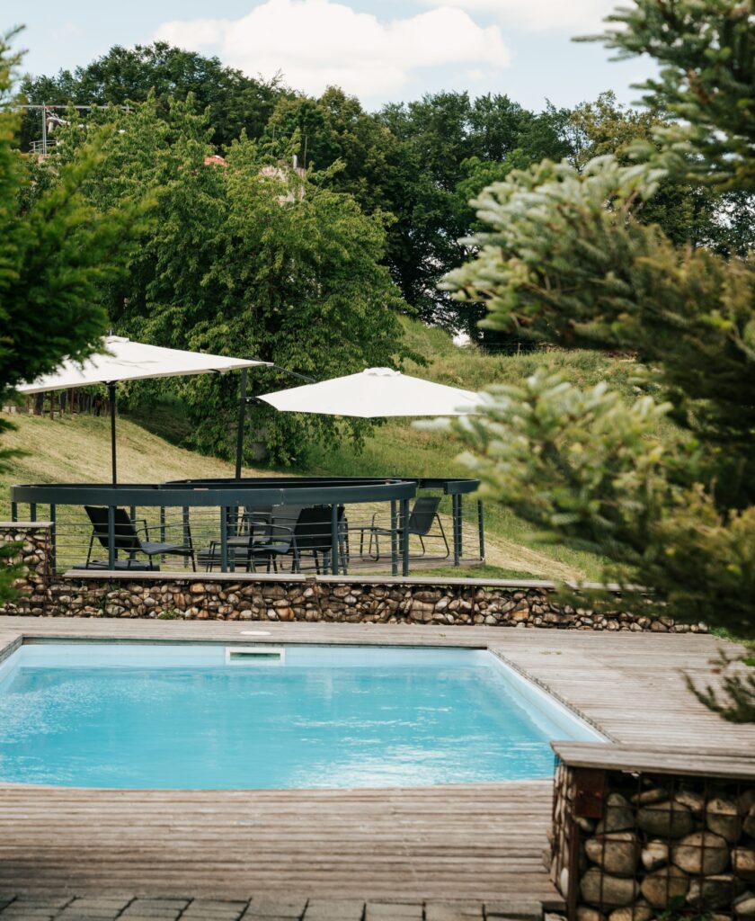 Rechteckiger Pool mit Holzdeck und Sitzbereich mit Tischen und weißen Sonnenschirmen vor bewaldetem Hang