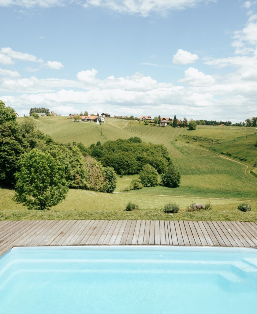 Blick über einen Pool mit Holzdeck auf eine hügelige Weinlandschaft mit grünen Wiesen, Bäumen und einem Dorf am Horizont unter blauem Himmel mit Wolken. Aussicht auf die besondere Ried Grubthal von Muster.Gamlitz.