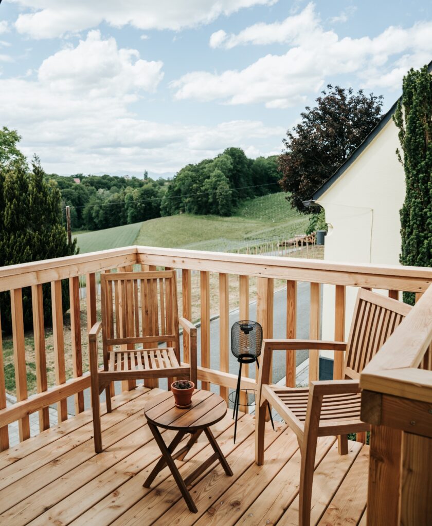 Balkon mit zwei Holzstühlen, kleinem runden Holztisch mit Blumentopf und Laterne, im Hintergrund grüne Hügel, Weinlagen und ein Haus