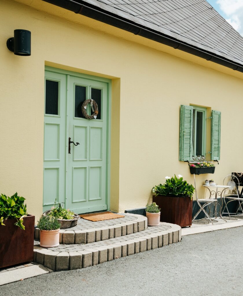 Eingang mit pastellgrüner Tür, Stufen aus grauen Steinen, zwei großen Pflanzkübeln und einem kleinen Sitzbereich mit Stuhl und Tisch vor einem gelben Haus