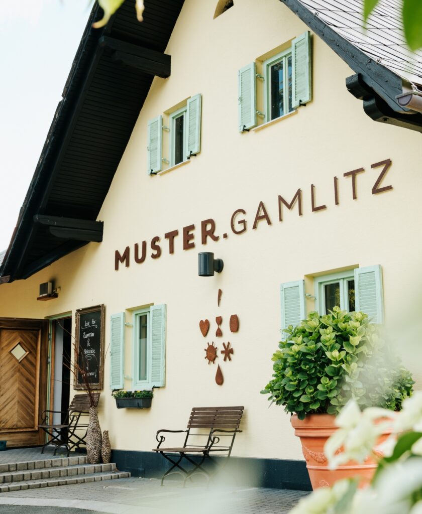 Fassade eines pastell-gelben Hauses mit der Aufschrift 'MUSTER.GAMLITZ', grünen Fensterläden, einer Bank und einem großen Blumentopf mit Pflanzen im Vordergrund