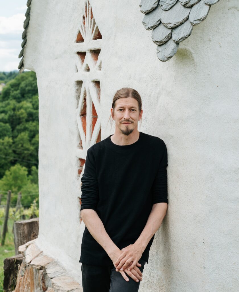Person – Reinhard Muster – mit schwarzem Langarmshirt lehnt an einer weißen Wand mit dekorativem Mauerwerk und Schindeldach im Freien. Direkt an der Ried Grubthal.