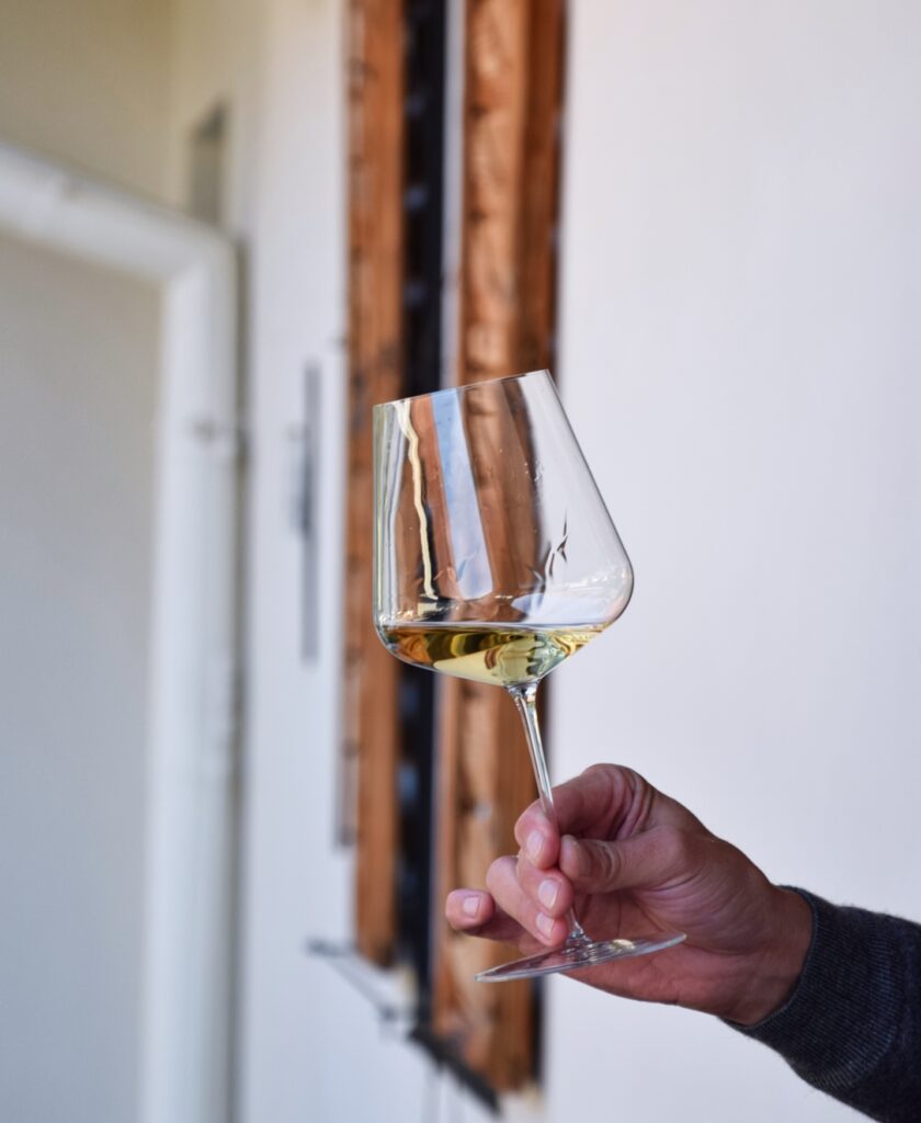 Hand hält ein Weinglas mit weißem Wein von Weingut muster.Gamlitz aus der Südsteiermark vor einem Fenster mit Holzrahmen