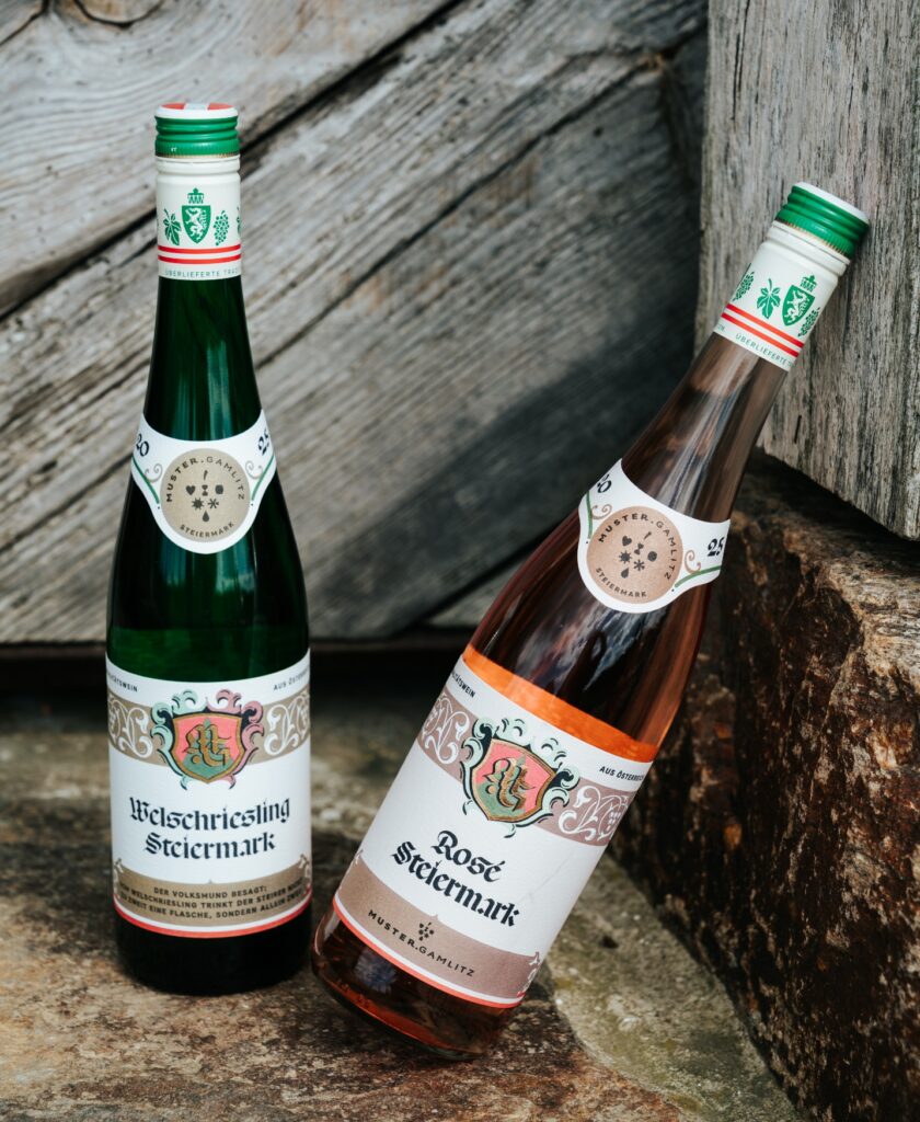 Zwei Weinflaschen mit Etiketten "Welschriesling Steiermark" und "Rosé Steiermark" lehnen an einer rustikalen Holz- und Steinoberfläche