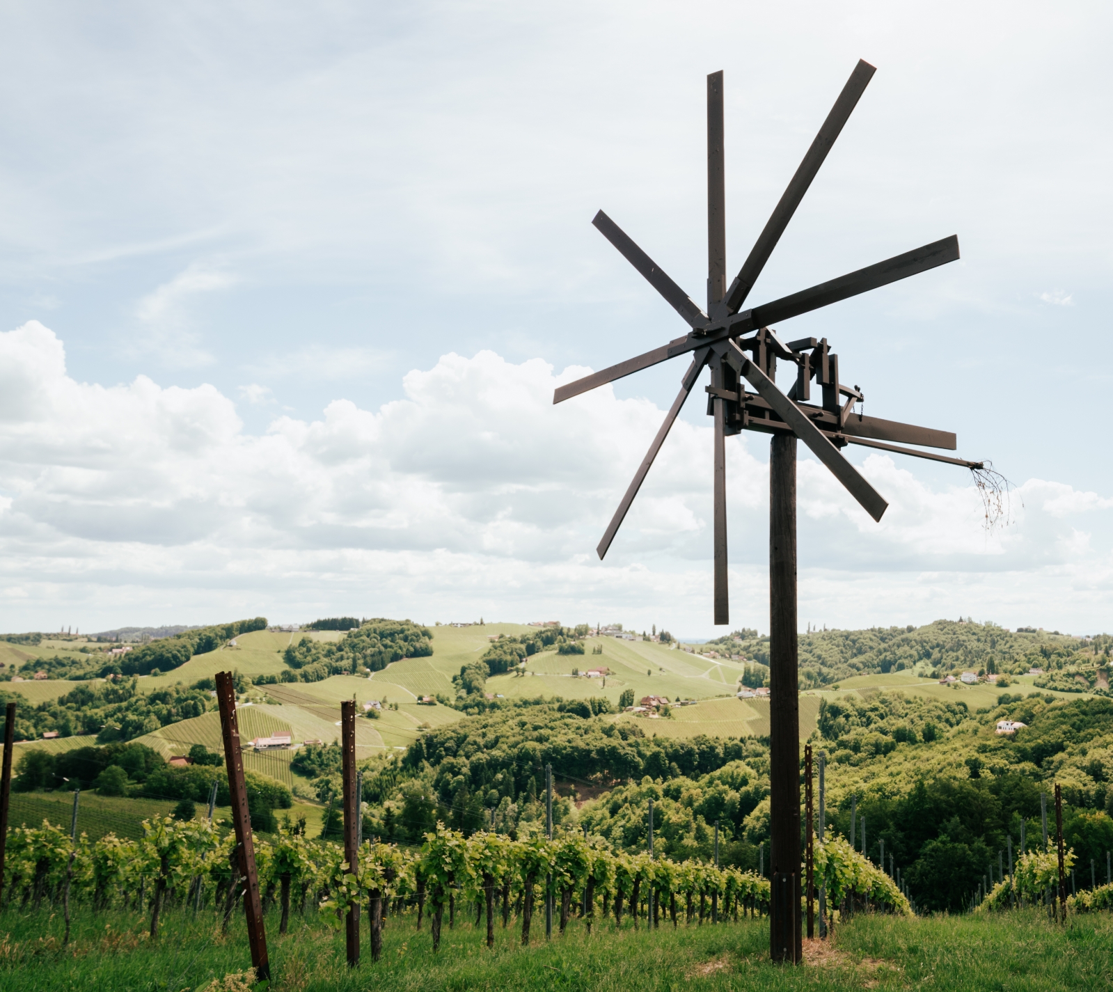 Metallskulptur mit radial angeordneten Balken in einer grünen Weinberglandschaft unter bewölktem Himmel