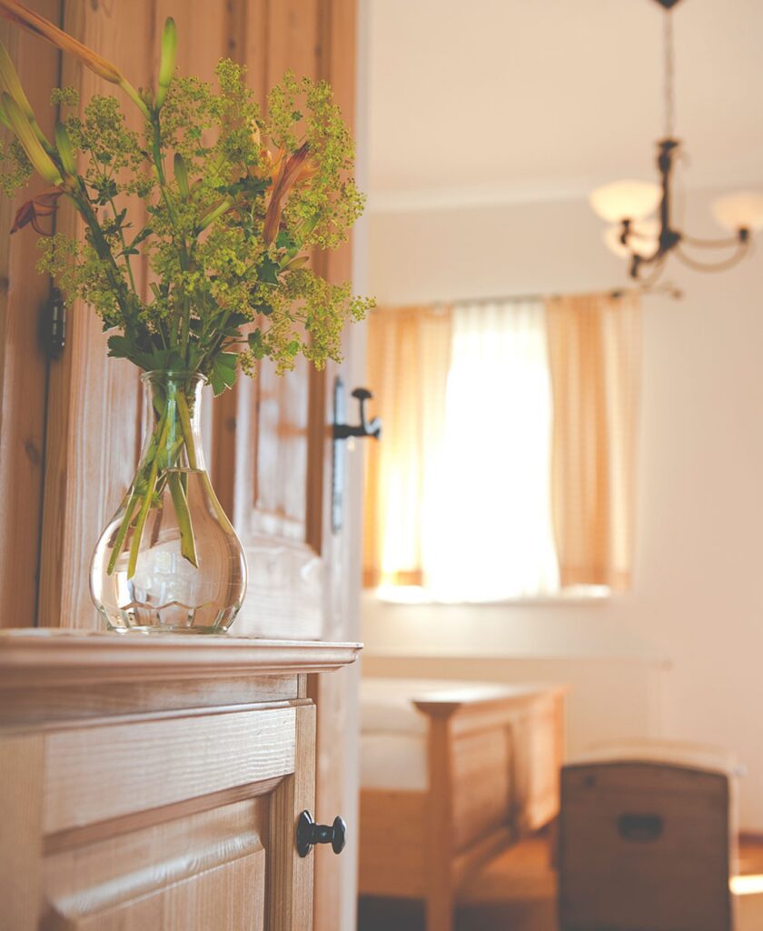 Vase mit grünen Zweigen auf einem Holztisch vor einem Fenster mit hellen Vorhängen und einem Holzbett im Hintergrund