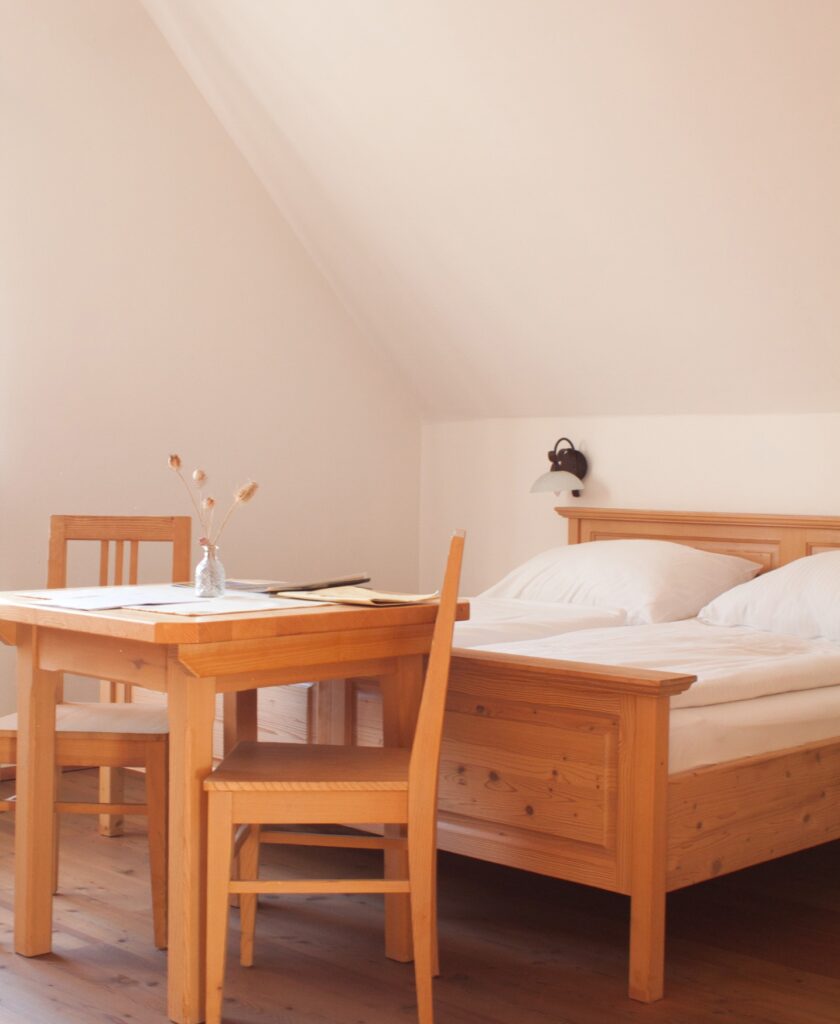 Holz-Doppelbett mit weißen Kissen und Decken neben einem Holztisch mit zwei Stühlen und einer Glasvase mit Trockenblumen in einem hellen Raum mit schräger Decke