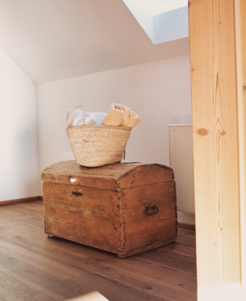 Alte hölzerne Truhe mit geflochtenem Korb und Decken darauf in einem hellen Raum mit Holzboden und Dachfenster