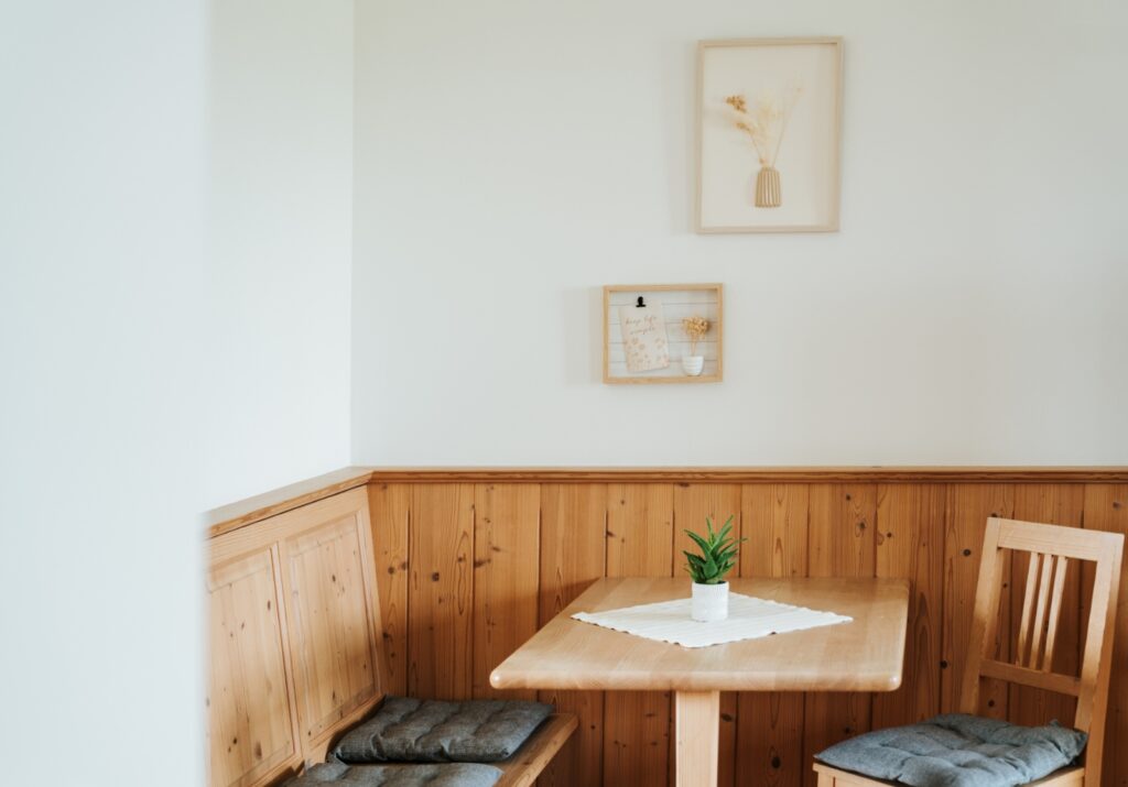 Holzecke mit Tisch, zwei Stühlen und Sitzbank mit grauen Sitzkissen, darüber zwei gerahmte Bilder an weißer Wand