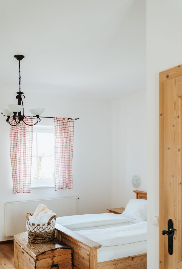 Schlicht eingerichtetes Schlafzimmer mit zwei Einzelbetten, Holzmöbeln, Korb mit Handtüchern auf einer Truhe und Fenster mit karierten Vorhängen
