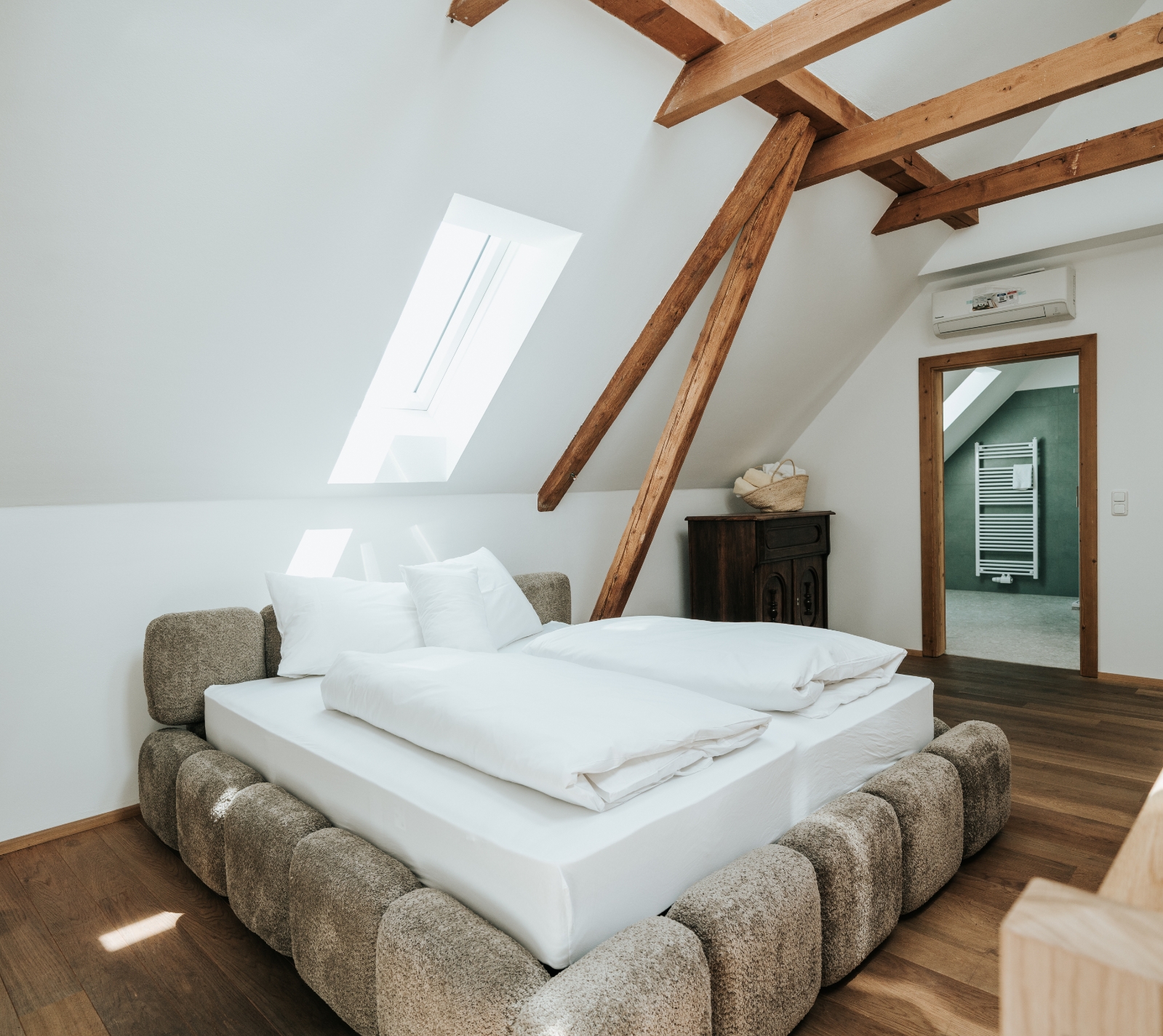 Schlafzimmer mit Dachschräge, Holzbalken, Bett mit gepolstertem Rahmen und weißer Bettwäsche, Holzboden und offenem Türrahmen zu einem angrenzenden Raum