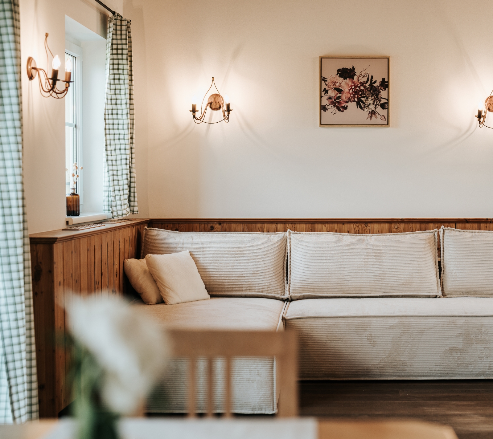 Helles Wohnzimmer mit hellbeigem Ecksofa, Holzwandverkleidung, zwei Wandlampen und einem Blumenbild an der Wand
