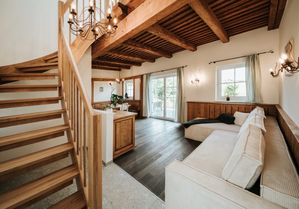 Wohnzimmer mit Holzbalkendecke, Treppe mit Holzgeländer, großen Fenstern und hellen Polstermöbeln