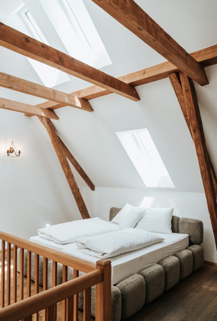 Schlafzimmer mit niedrigem, schrägem Dach, sichtbaren Holzbalken, zwei Dachfenstern und einem gepolsterten Bett mit weißer Bettwäsche
