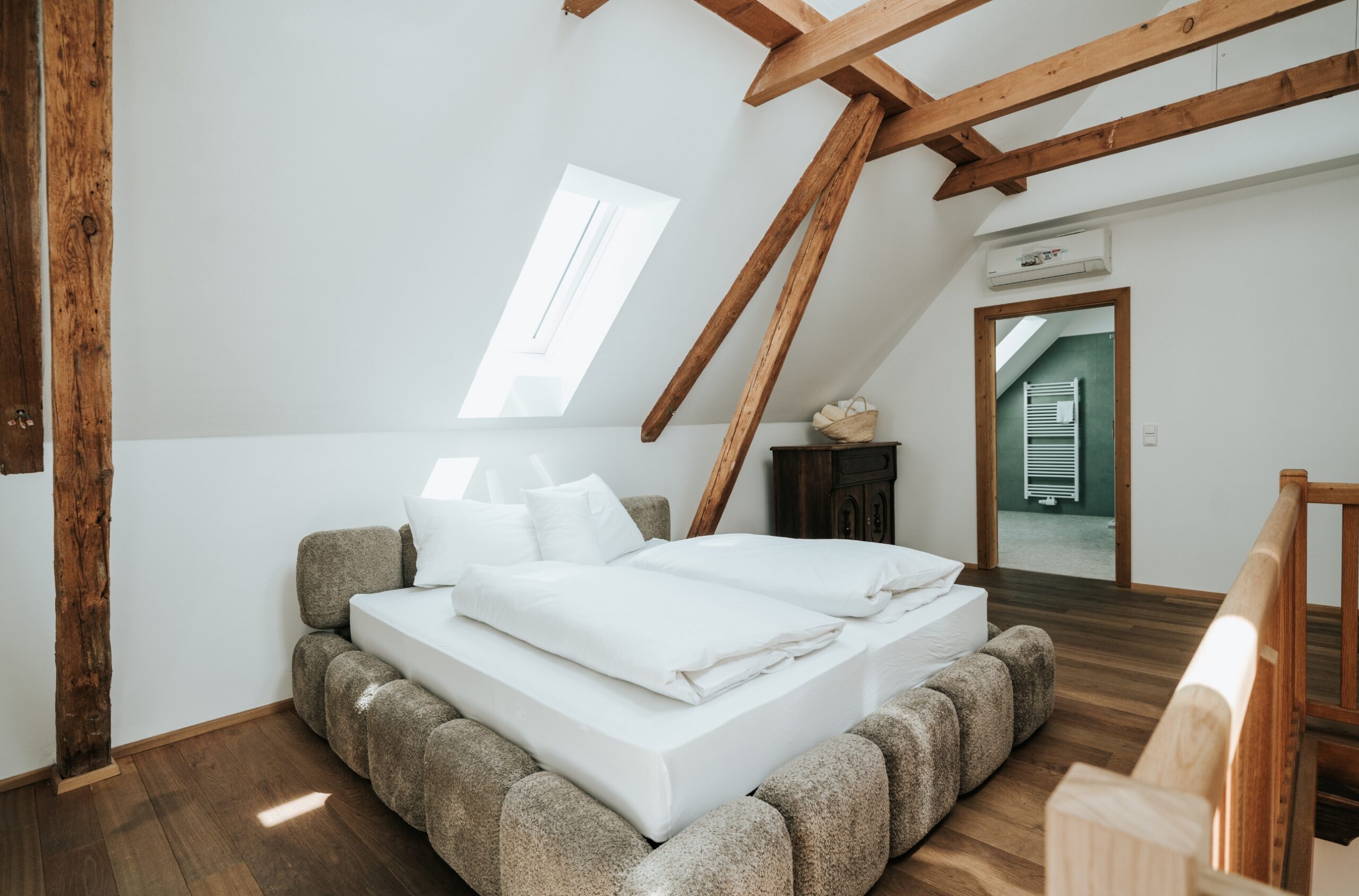 Schlafzimmer mit Dachschräge, Holzbalken, Bett mit gepolstertem Rahmen und weißer Bettwäsche, Holzboden und offenem Türrahmen zu einem angrenzenden Raum