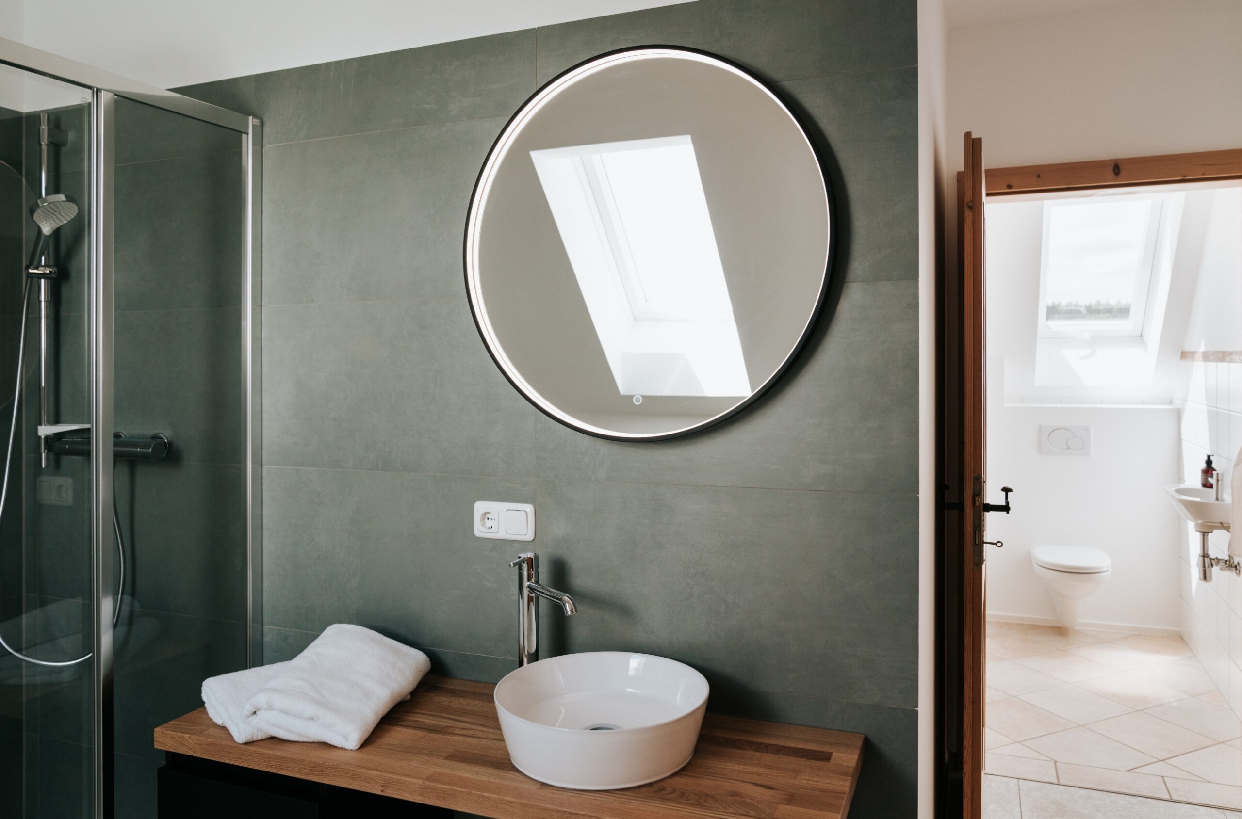 Modernes Badezimmer mit rundem Spiegel, weißem Waschbecken auf Holzablage und Handtüchern, Blick in angrenzenden Raum mit Toilette und Fenster