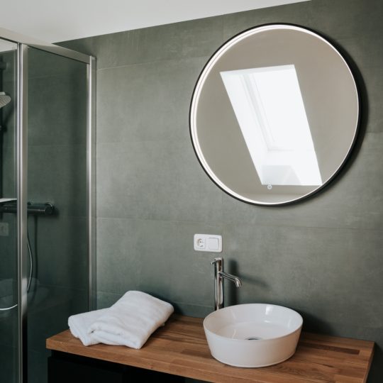 Rundes Wandspiegel über einem Holzwaschtisch mit rundem Aufsatzwaschbecken und Handtüchern neben einer Glasdusche in einem modernen Badezimmer